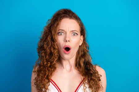 Young woman with red curly hair surprised expression open mouth stands against vivid blue background wearing a white red topの写真素材