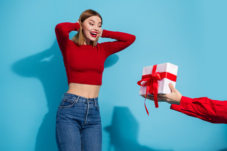 Excited young woman receiving a beautifully wrapped gift, expressing joy and surprise against a vibrant blue backgroundの写真素材