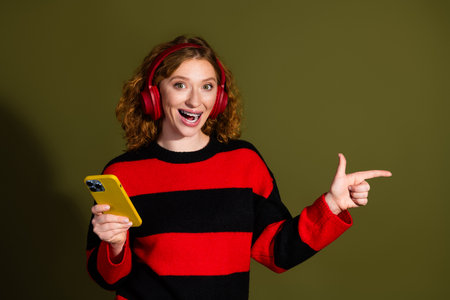 Young girl with red hair wearing a red and black striped sweater listens to music with red...