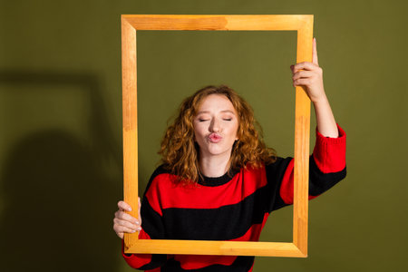 Playful young woman with curly red hair holds a wooden frame and puckers her lips against a green backdropの写真素材