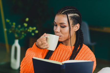 Relaxed moment at home a young woman with braids sipping coffee and reading a book in a cozy orange...