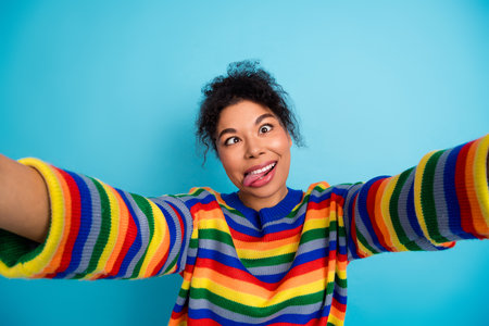 Young woman with rainbow striped sweater smiles and stretches toward the camera against a blue backgroundの写真素材