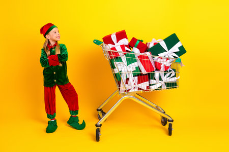 Festive young elf dressed in vibrant costume posing near cart of gift boxes in cheerful yellow ambiance celebrating holidaysの写真素材