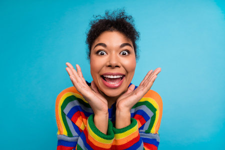 Joyful young woman with curly hair wearing sweater smiles with hands framing her face blue background perfect for lifestyle advertising fashionの写真素材