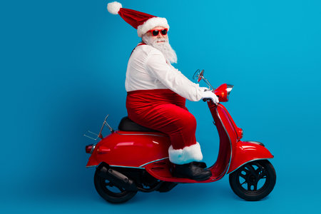 Santa on a red scooter wearing hat beard costume and white gloves in a bright blue studio for Christmas vibeの写真素材