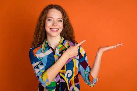 Photo of lovely adorable cheerful girl wear trendy print clothes hold banner news empty space isolated on vivid orange color backgroundの写真素材