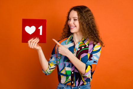 Photo of charming adorable girl blogger wear trendy print clothes hands hold red heart symbol isolated on vivid orange color backgroundの写真素材