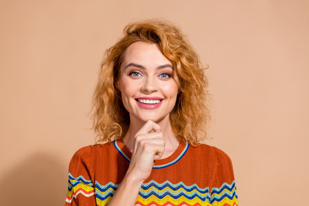 Portrait of pleasant girl with ginger hairdo wear knit t-shirt smiling hold fingers on chin isolated on beige color backgroundの写真素材