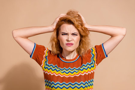 Portrait of stressed unhappy girl with ginger hairdo wear knit t-shirt hold hands on head isolated on beige color backgroundの写真素材