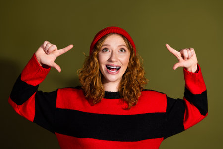 Young woman with red hair in red and black striped sweater smiles and points with both hands in a playful fashion lifestyleの写真素材