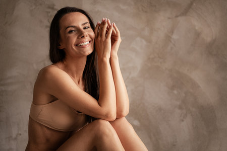Portrait of attractive brunette hair young woman no retouch in underwear palms together sitting floor isolated over gray color backgroundの写真素材