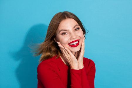 Beautiful Woman in Red Top Smiling Confidently Against Vibrant Blue Background with Hands on Face, Expressing Happinessの写真素材
