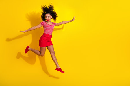 Cheerful young woman in a vibrant outfit jumping with joy against a colorful yellow background,...