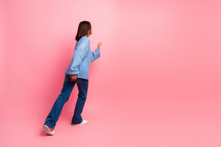 Young woman in blue knit sweater walks forward against a pink background showcasing casual style and modern fashionの写真素材