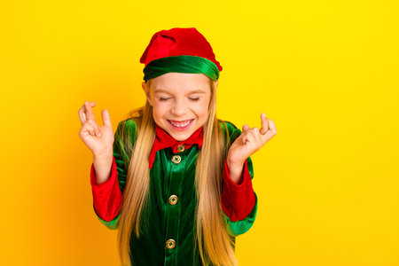 Smiling young girl dressed as a festive elf with crossed fingers in a colorful Christmas theme, yellow backgroundの写真素材