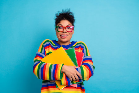 Colorful student woman with striped sweater holding notebooks smiles against blue background for fashion lifestyle shoot showcasing diversityの写真素材