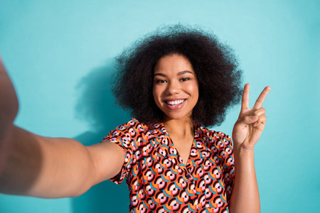 Young cheerful woman with natural hair taking a selfie wearing a vibrant geometric shirt against a bright blue background smiling and showing a peace signの写真素材
