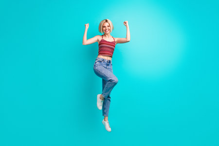 Young woman jumping with joy in stylish casual outfit against bright teal background for fitness...