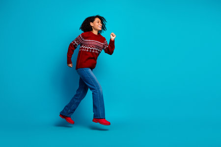 Joyful young woman in a red christmas sweater jumps with energy against a blue background celebrating the holiday seasonの写真素材