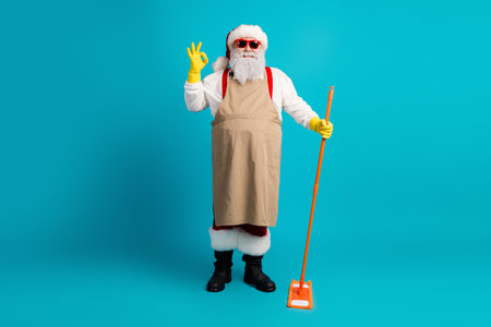 Santa in beige apron and yellow gloves with orange mop stands against blue background for festive season shopping and holiday celebrationの写真素材