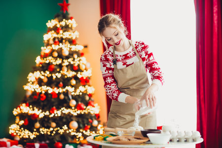Playful home scene with a smiling woman in an apron baking beside a glowing Christmas tree full of lights and festive decorations at homeの写真素材