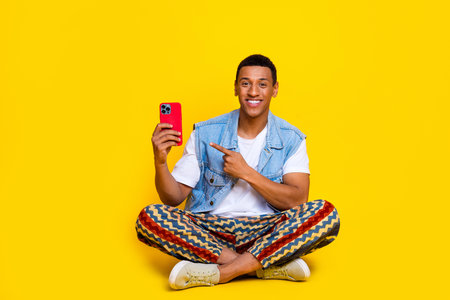 Full length photo of cheerful nice guy dressed jeans waistcoat sit indicating at smartphone in arm isolated on yellow color backgroundの写真素材