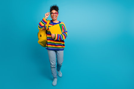 Bright multicolor striped sweater student smiles wearing yellow backpack with notebooks against blue backgroundの写真素材