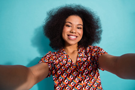 Young woman smiling while taking a selfie in a colorful geometric shirt against a blue backgroundの写真素材