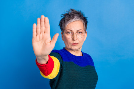 Photo of serious elderly woman wear stylish clothes stop symbol isolated on blue color backgroundの写真素材