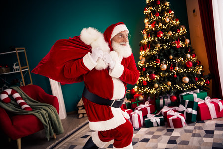 Santa carries a large sack through a cozy home beside a lit Christmas tree filled with gifts and festive lights creating a warm holiday sceneの写真素材