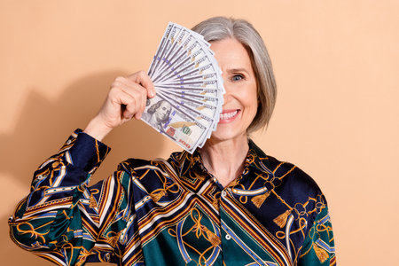 Photo of satisfied retired person with bob hairdo wear print blouse holding dollars isolated on beige color backgroundの写真素材