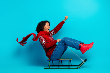 Playful young woman on a sled jumps for joy in a red sweater and scarf against a bright blue...