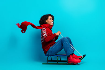 Bright multiethnic woman in red sweater and scarf rides a small sled against a blue background creating a playful festive winter sceneの写真素材