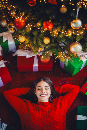 Funny dreamy christmas moment as a young woman in red sweater rests under a lit christmas tree with gifts and warm glowの写真素材