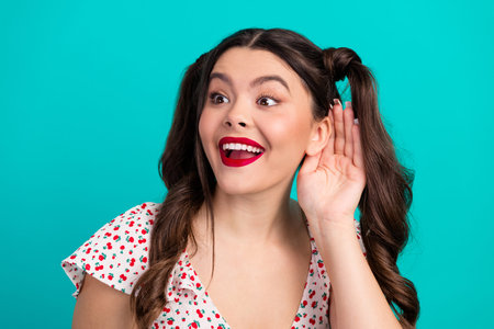 A glamorous young woman with brunette ponytails wears a cherry print top, smiles widely, and cups...