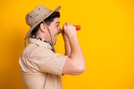 A cheerful young man in safari style uniform peers through a small red telescope against a bold...