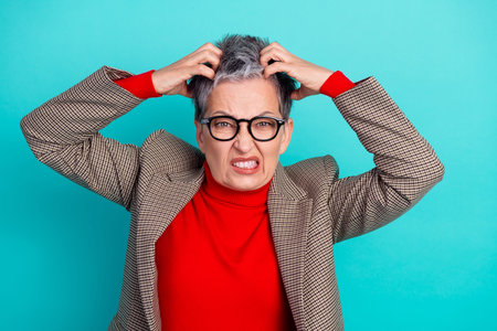 Aged businesswoman with short gray hair and glasses pulls her hair in a tense moment, wearing a red...