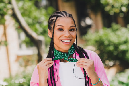 A young woman with braids ties a green bandana around her neck while standing in a sunny garden...