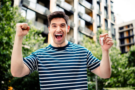 A cheerful man proudly displays keys outdoors in a vibrant urban setting, celebrating success and joy against the backdrop of modern apartments.