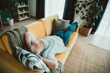 A senior man with a white beard lies on a yellow couch, legs up, in a sunny living room. Casual home setting conveys comfort, relaxation, and domestic leisure for real estate and interior design themes.