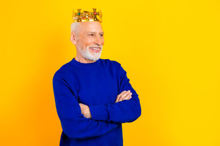 A cheerful elderly man in a blue sweater wears a gold crown and smiles with folded arms against a vivid yellow backdrop, conveying confidence, playfulness, dignity and celebratory mood.