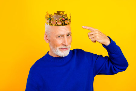 Playful senior man portrait wearing a gold crown and blue sweater, pointing to his head with a confident, amused expression on a bright yellow background. Concept of royalty, confidence and humor.
