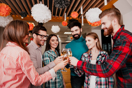 A diverse group of young adults clink glasses and smile at a decorated office party, capturing a warm moment of celebration, teamwork and friendship in a relaxed indoor setting.