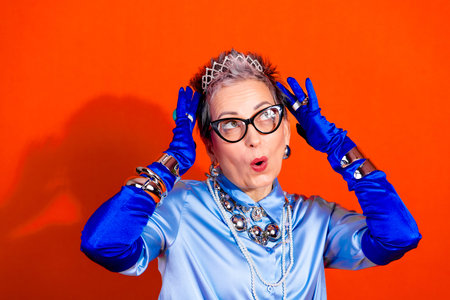 A stylish mature woman with gray short hair, black glasses, and layered jewelry expresses surprise and playfulness while wearing a satin blue outfit. Bright orange backdrop adds bold fashion vibes and energy.