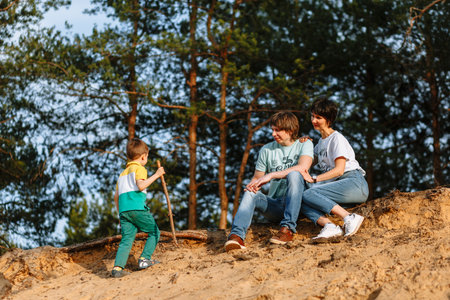 A child with a stick climbs up the sand to the sitting father and motherの写真素材