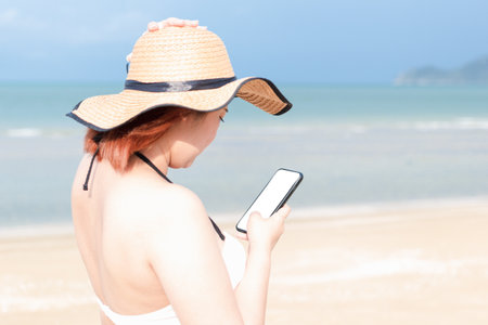 A woman wearing a swimming suit and a sunhat is operating a mobile phone standing by the beach. Blank cell phone for mock up.の写真素材