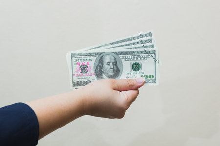 The hand of a white female adult in a blue shirt is holding three hundred dollar bills on a gray background.の写真素材