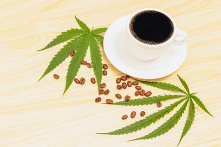 Hot black coffee in a cup with a white saucer and coffee beans sprinkled on a wooden table and decorated with hemp leaves, cannabis coffee relaxing drink concept.の写真素材