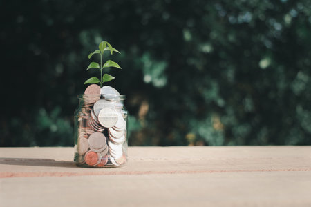 Plant growing in coin in glass jar placed on wooden floor and green blur background, money saving and investment idea.の写真素材