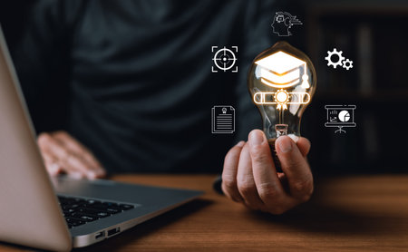 A person holds a glowing light bulb with icons of graduation,charts,documents,and gears symbolizing...